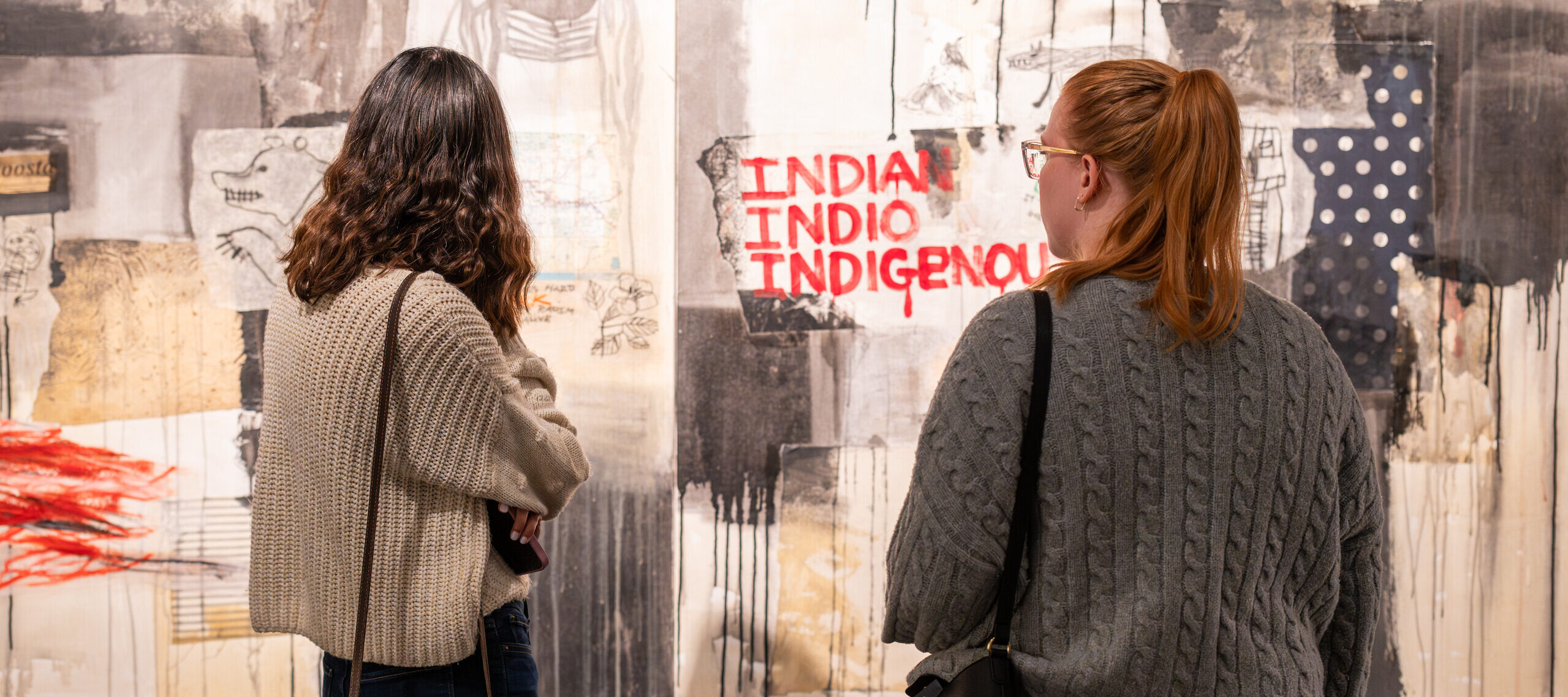 Two female visitors in grey sweaters look at an abstract painting. The painting is primarily beige and grey blocks of color, and the words "Indian" "Indio" and "Indigenous" are written in red paint.