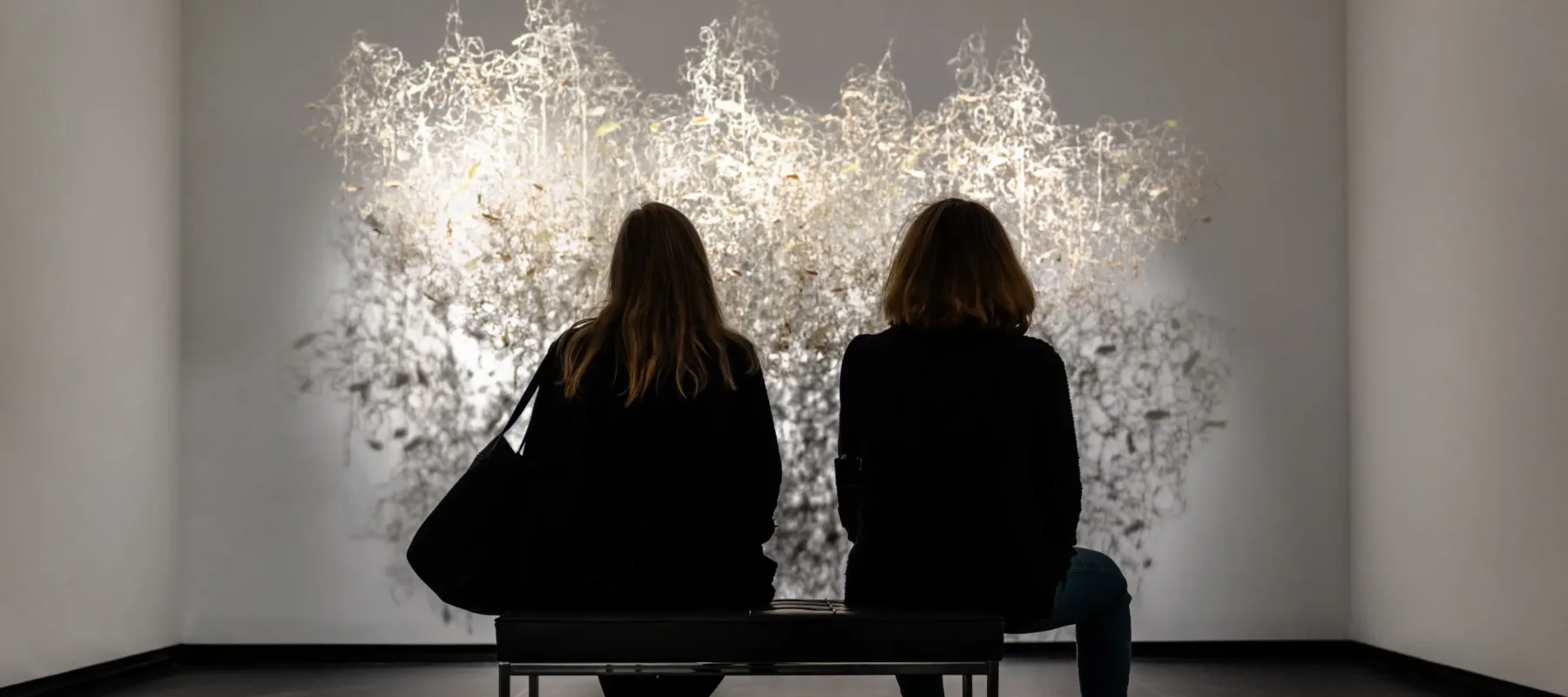 Two museum visitors sit on a bench and observe a brightly lit artwork in a modern gallery.