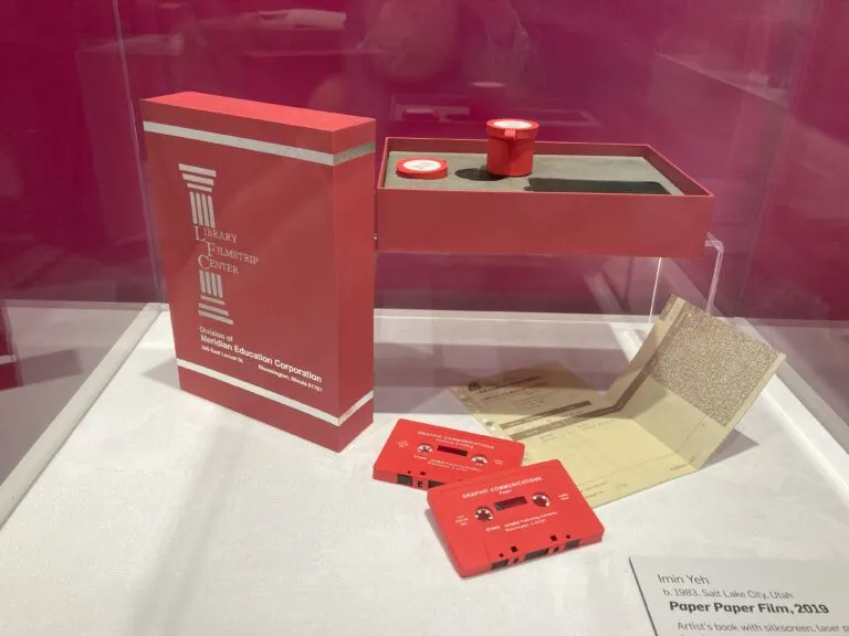 Two red cassette tapes and a printed receipt accompanied by two red film canisters and a gray book the size of a cassette tape with the title "Paper Paper Film" embossed on its cover in a red storage box with a lid that reads "Education Corporation."