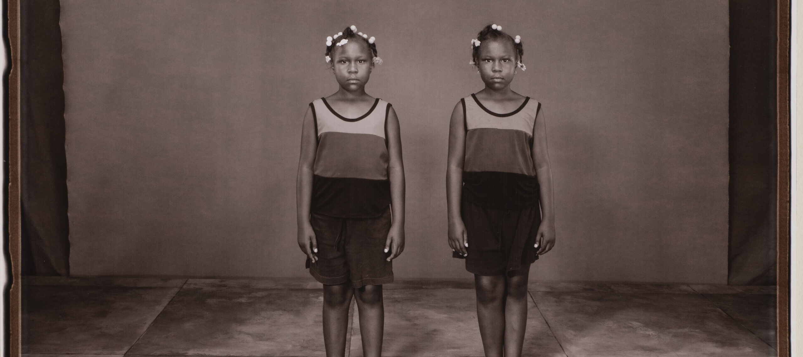 A photograph of two twin adolescents with dark skin tone wearing matching clothing, hairstyles, and poses.
