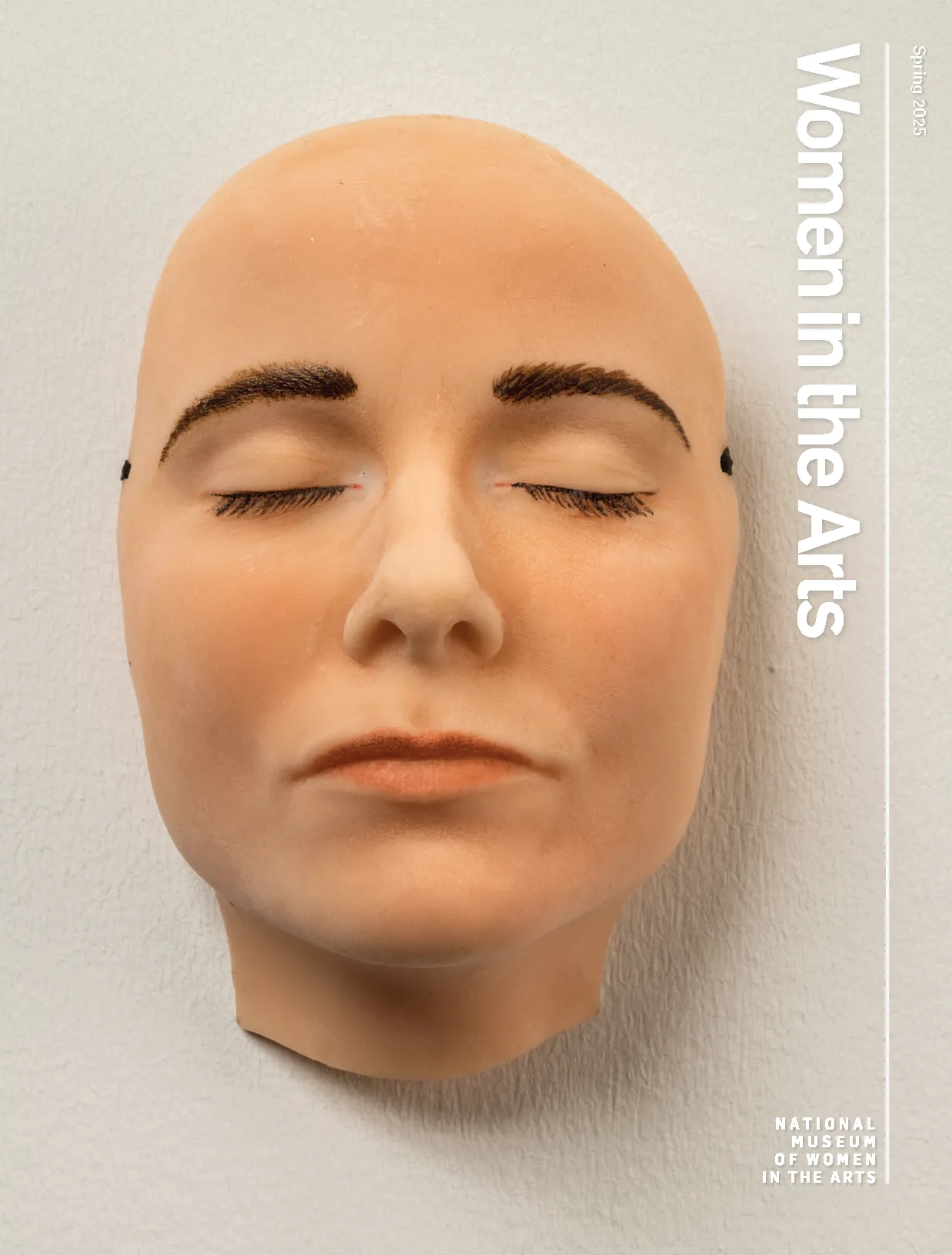 The cover of Women in the Arts Spring 2025 issue with an image of a highly realistic mask of a light-skinned face with dark brown eyebrows and eyelashes that appears to be sleeping—its eyes and mouth are closed and relaxed. The mask hangs on a white wall.