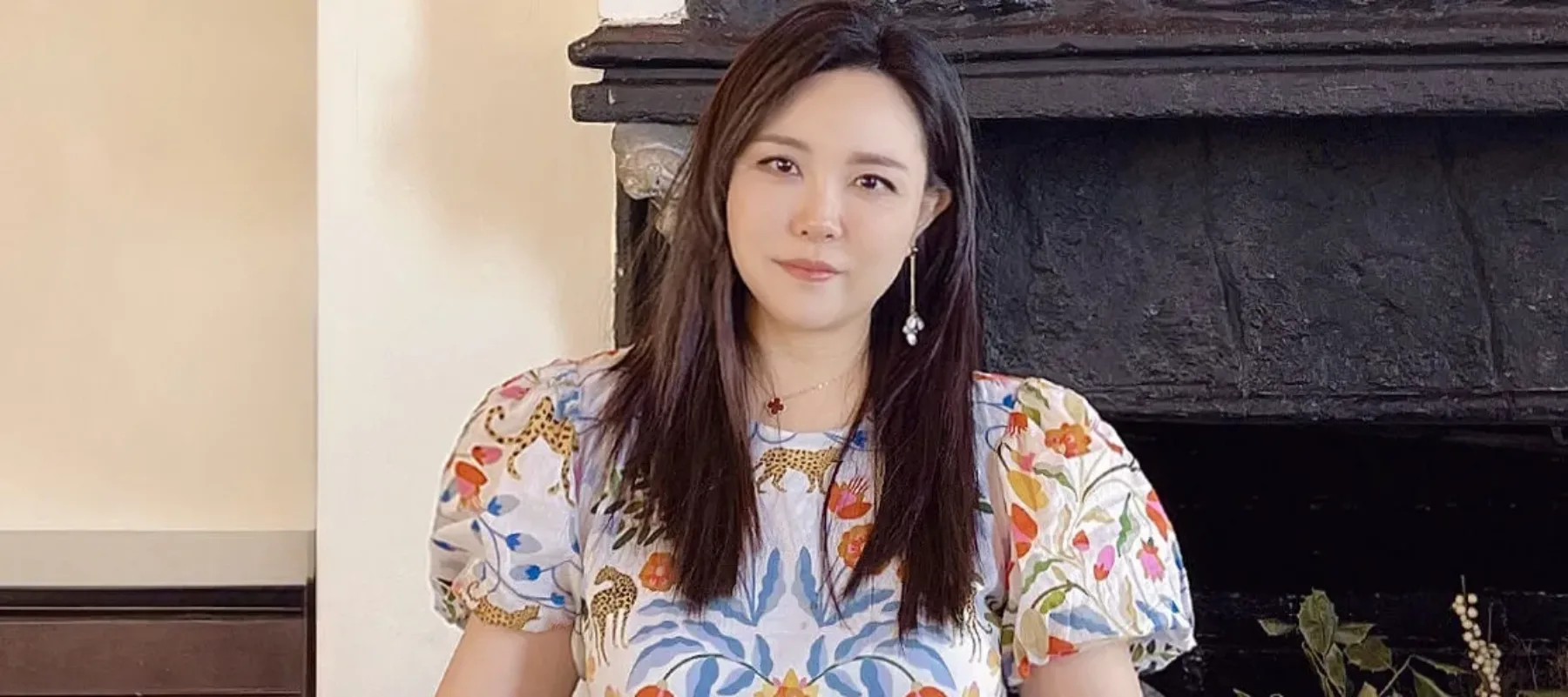 A light-skinned Asian woman stands behind three large decorative boxes featuring a floral motif. She wears a puffy-sleeved blouse in the same pattern. She looks at the camera with a neutral expression. Behind her is a large stone fireplace and in front of her are colorful patterned scarves folded and overlapping.