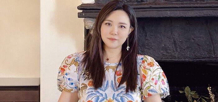 A light-skinned Asian woman stands behind three large decorative boxes featuring a floral motif. She wears a puffy-sleeved blouse in the same pattern. She looks at the camera with a neutral expression. Behind her is a large stone fireplace and in front of her are colorful patterned scarves folded and overlapping.