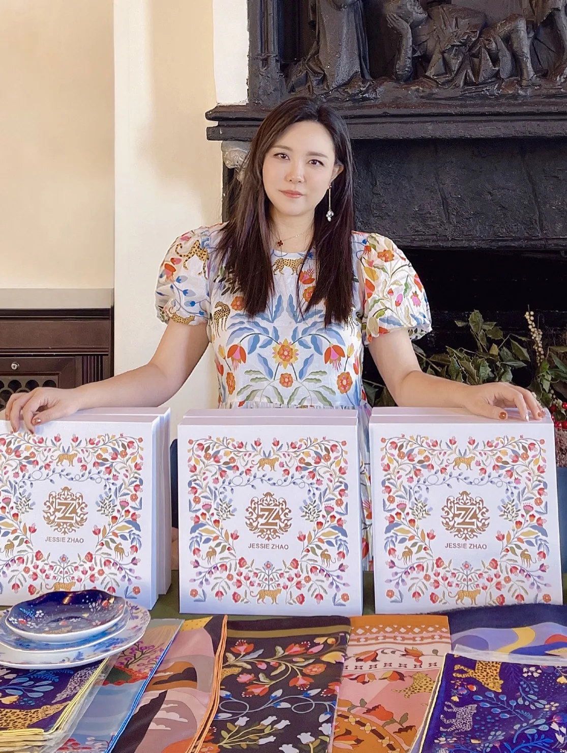 A light-skinned Asian woman stands behind three large decorative boxes featuring a floral motif. She wears a puffy-sleeved blouse in the same pattern. She looks at the camera with a neutral expression. Behind her is a large stone fireplace and in front of her are colorful patterned scarves folded and overlapping.