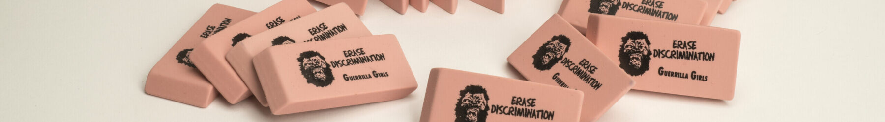 A pile of pink pencil erasers each with a screen print of a growling gorilla face mask, the command “ERASE DISCRIMINATION” in all capital letters, and the phrase “Guerrilla Girls,” which is the name of the troop of anonymous artists responsible for this work.