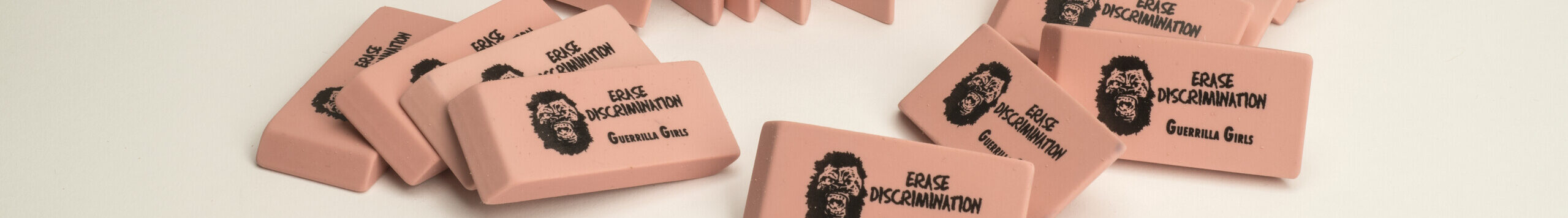 A pile of pink pencil erasers each with a screen print of a growling gorilla face mask, the command “ERASE DISCRIMINATION” in all capital letters, and the phrase “Guerrilla Girls,” which is the name of the troop of anonymous artists responsible for this work.