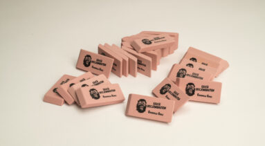 A pile of pink pencil erasers each with a screen print of a growling gorilla face mask, the command “ERASE DISCRIMINATION” in all capital letters, and the phrase “Guerrilla Girls,” which is the name of the troop of anonymous artists responsible for this work.
