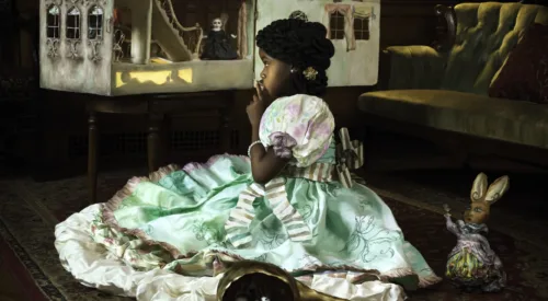 A young girl with medium-dark skin tone wears an elaborate dress and sits on a rug in a library, gazing at a dollhouse with lit windows. An empty gourd filled with cotton bolls and a rabbit toy are nearby. The dollhouse harbors figures, one of whom appears to be dressed as a Confederate soldier.
