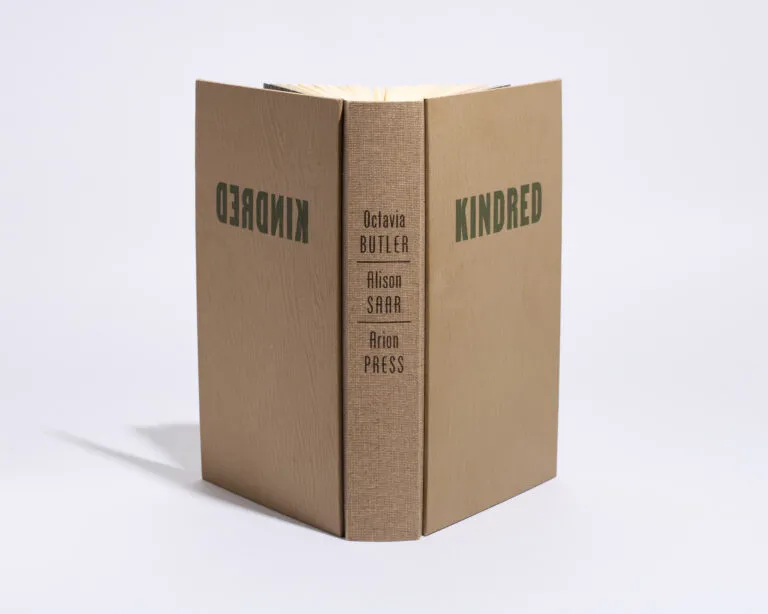 A copy of the book “Kindred” stands straight against an all-white background. The spine reads Octavia Butler (the author), Alison Saar (the artist who created the art book), and Arion Press (the press).