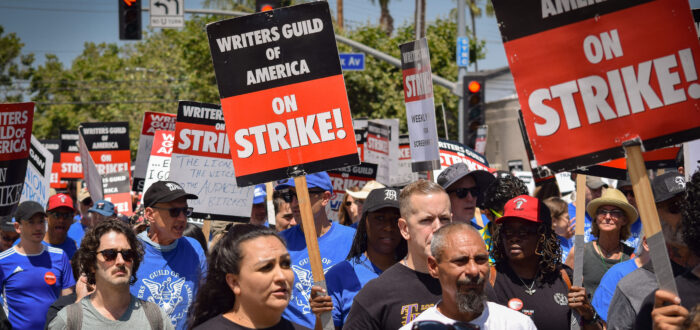 A large group of people diverse in age, race, and gender, hold picket signs and appear to be marching down a street. The signs say "Writer's Guild of America" on STRIKE!