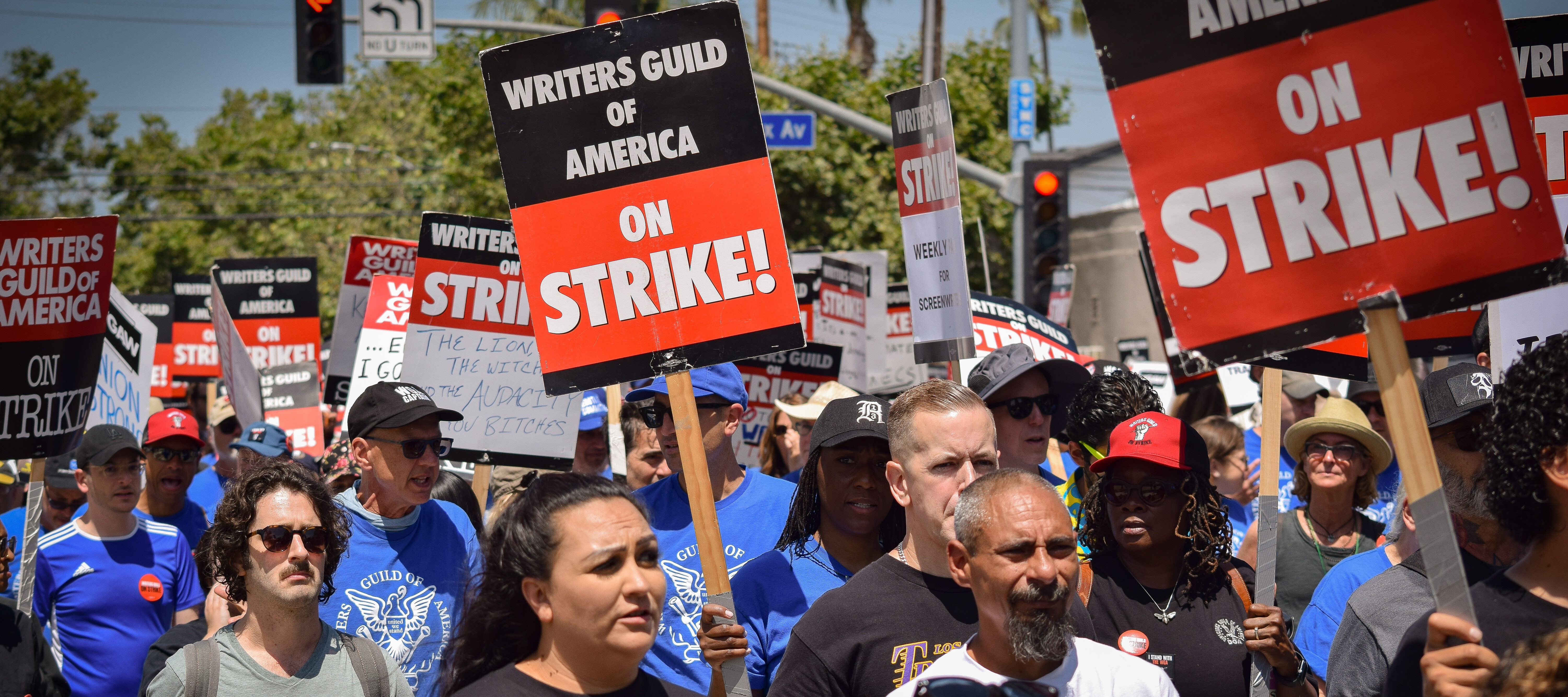 A large group of people diverse in age, race, and gender, hold picket signs and appear to be marching down a street. The signs say "Writer's Guild of America" on STRIKE!