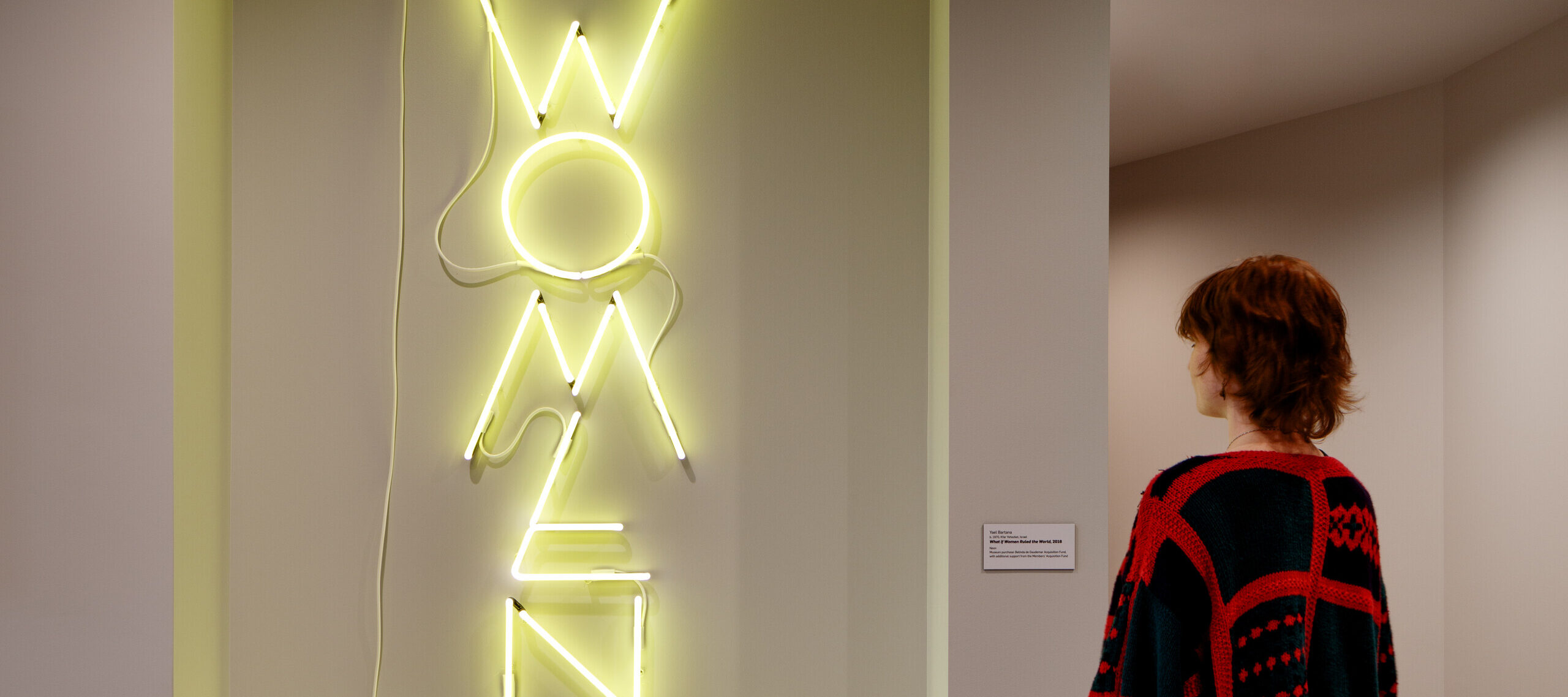 A museum visitor looks at a neon artwork hanging on a wall. The phrase "What if Women Ruled the World" appears sculpted with neon yellow tube lights, in all capital letters. The letters of “WOMEN” are larger than the rest and arranged vertically.
