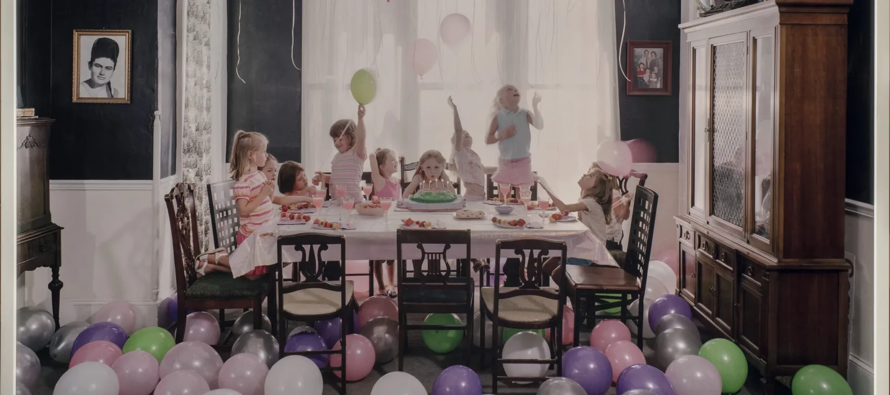 Green, purple, silver, white and pink balloons litter the floor and cover the ceiling of a navy-walled family dining room. In the middle of the room, a table is set with a pink table cloth. In the center of the table, a young girl blows out the candles on her birthday cake while looking straight ahead. Around her, nine other young girls jump, spin, and play.
