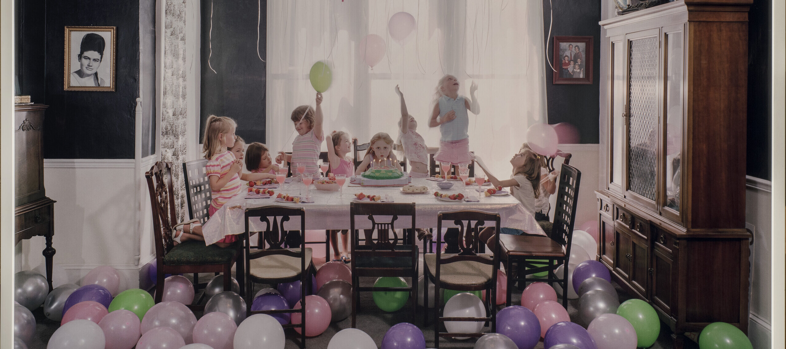 Green, purple, silver, white and pink balloons litter the floor and cover the ceiling of a navy-walled family dining room. In the middle of the room, a table is set with a pink table cloth. In the center of the table, a young girl blows out the candles on her birthday cake while looking straight ahead. Around her, nine other young girls jump, spin, and play.