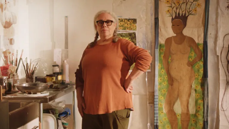 A woman with medium-light skin tone, gray pigtails, and large black glasses stands confidently in an art studio, wearing an orange sweater. Behind her is a large painting of a standing figure with flowers for hair.