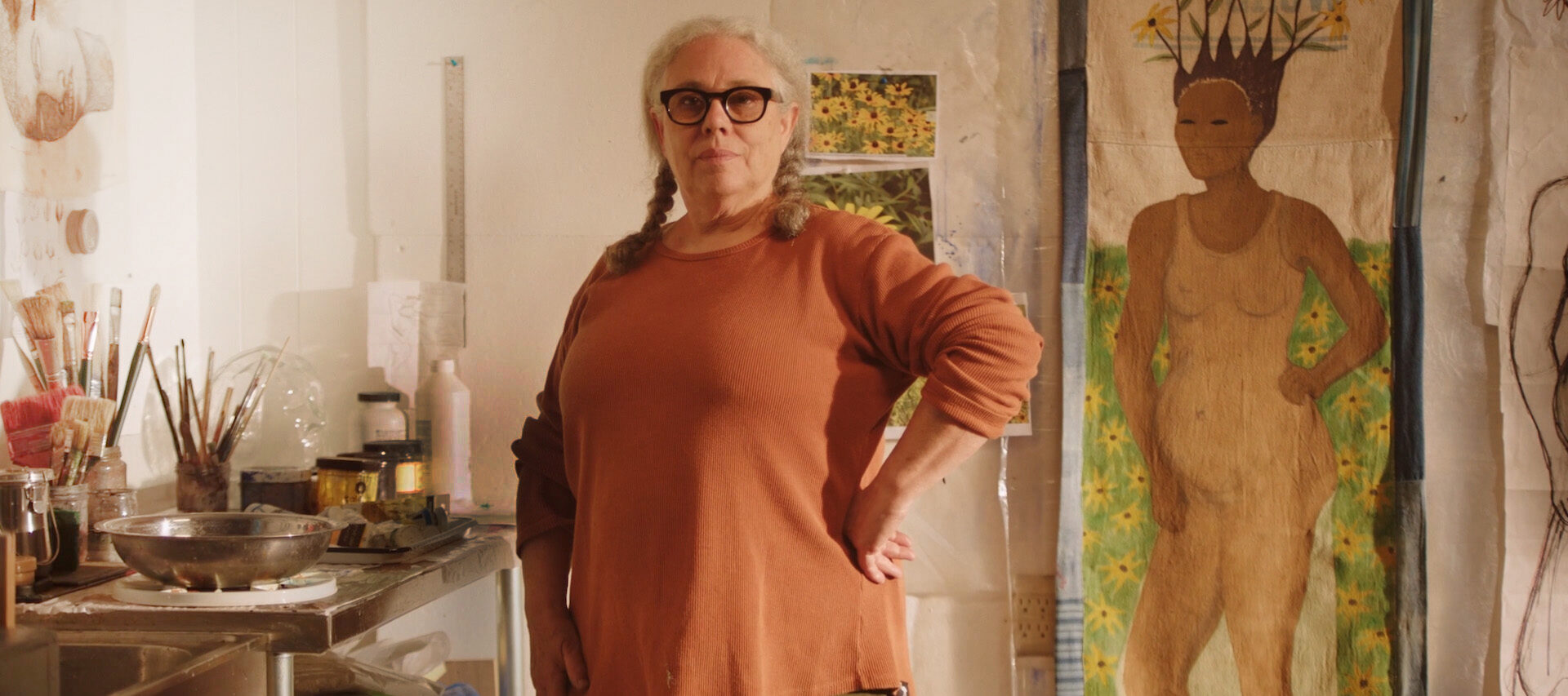 A woman with medium-light skin tone, gray pigtails, and large black glasses stands confidently in an art studio, wearing an orange sweater. Behind her is a large painting of a standing figure with flowers for hair.