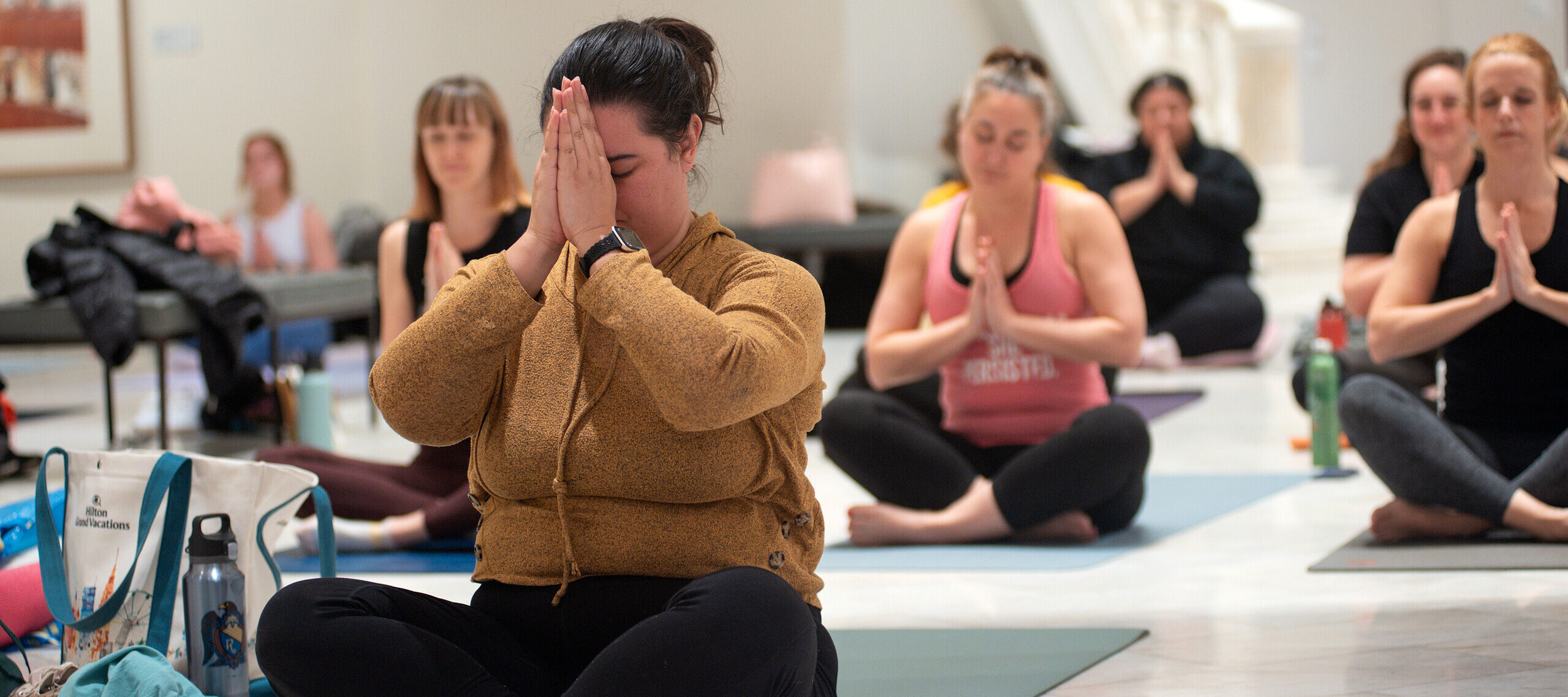 A crowd participates in a yoga class in the Great Hall of the National Museum of Women in the Arts. They all wear athletic apparel and sit cross-legged on their own yoga mats, with their hands touching at their chests.