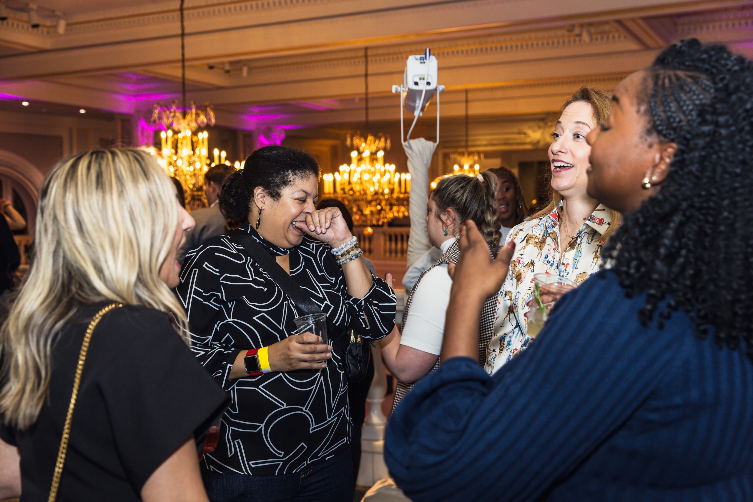A group of people are socializing and laughing in an elegant room with chandeliers. They appear to be enjoying a lively conversation. One person holds a drink, and the atmosphere seems joyful and relaxed.