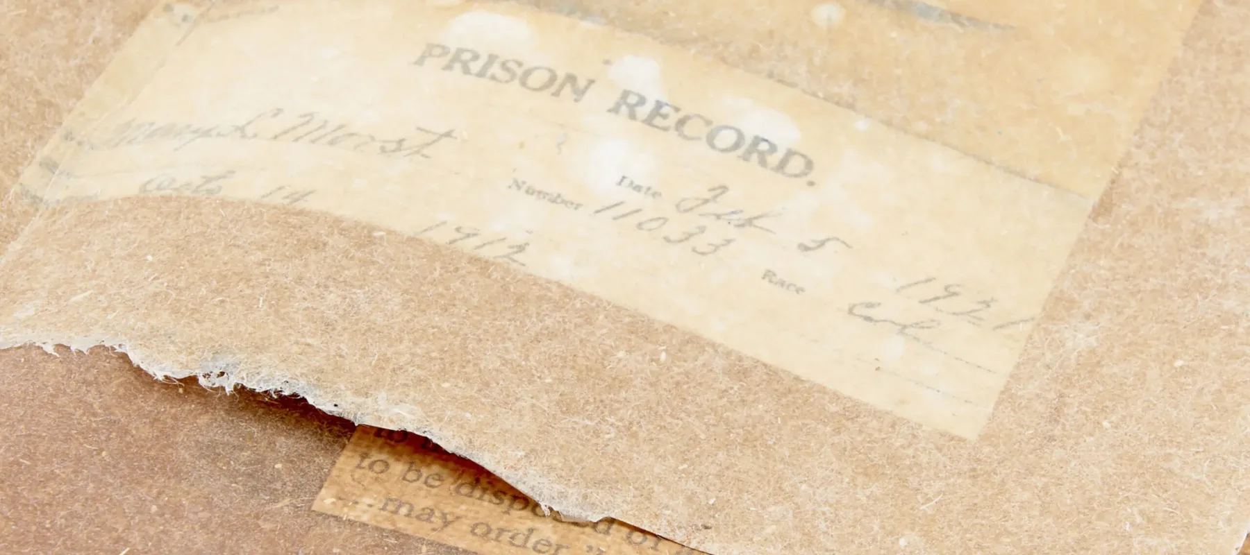 A  detailed view of a box-shaped book featuring aged, hand cut paper, layered with torn edges. The words "prison record" can be seen through the semi-translucent pages.