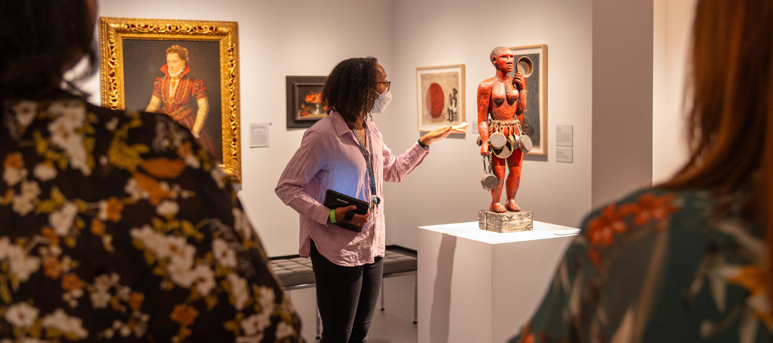 A woman with dark colored skin, a lavender button down shirt, and black jeans, looks and points at a red sculpture of a nude figure with a skirt of puts and pans, displayed on a white pedestal.