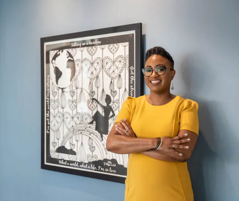 A young, dark-skinned woman with short, curly brown stands wearing a bright yellow outfit. She smiles directly to the camera with casually crossed arms standing next to an artwork in the background.