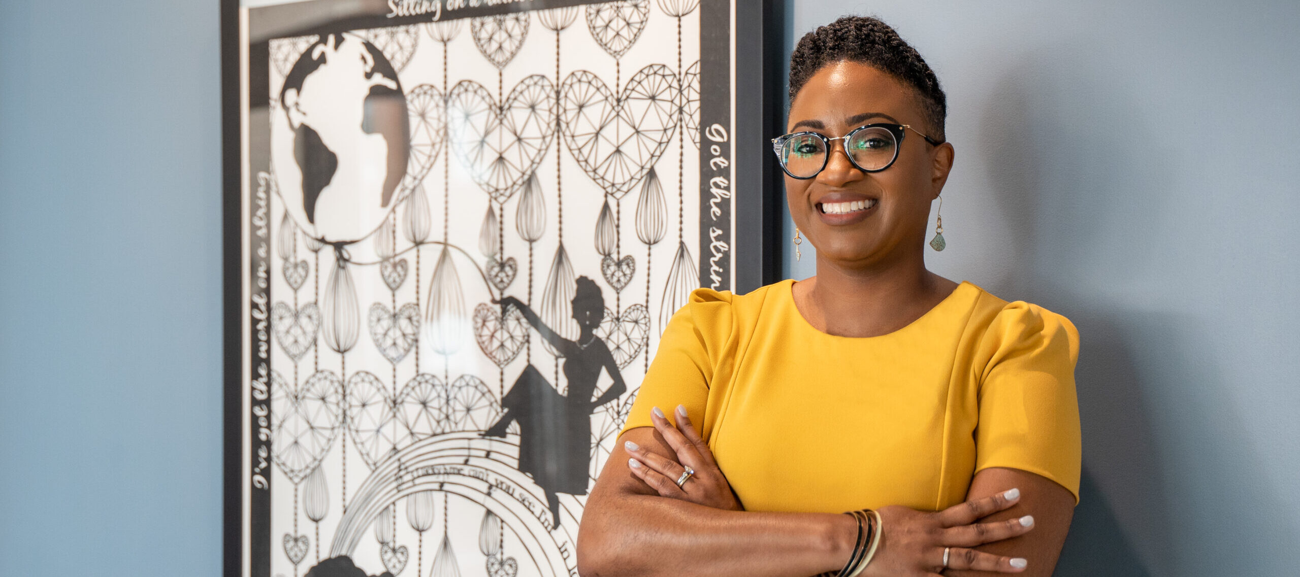 A young, dark-skinned woman with short, curly brown stands wearing a bright yellow outfit. She smiles directly to the camera with casually crossed arms standing next to an artwork in the background.