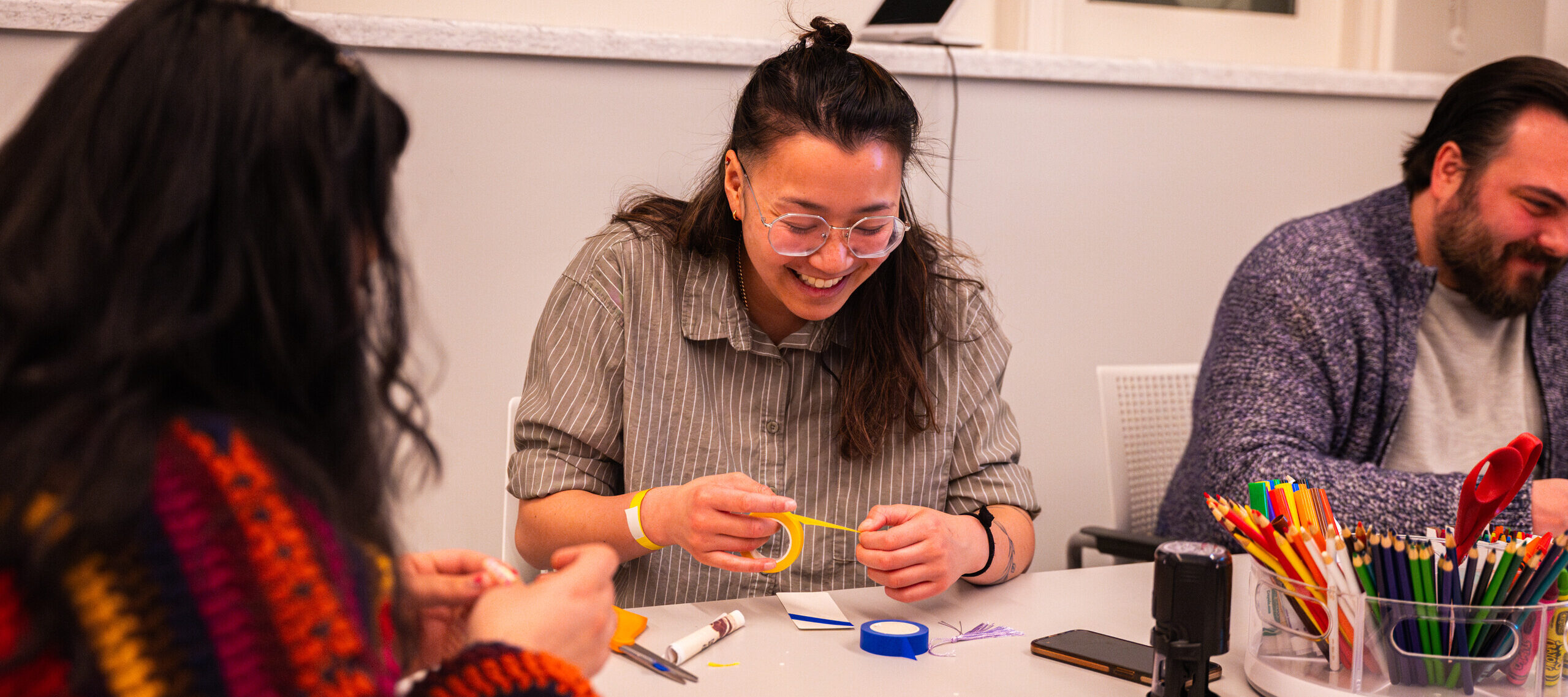 Three people sit at a grey table, decorating paper bookmarks. In the middle, a woman with light-medium skin, brown hair, and clear glasses laughs as she pulls yellow tape over her bookmark.