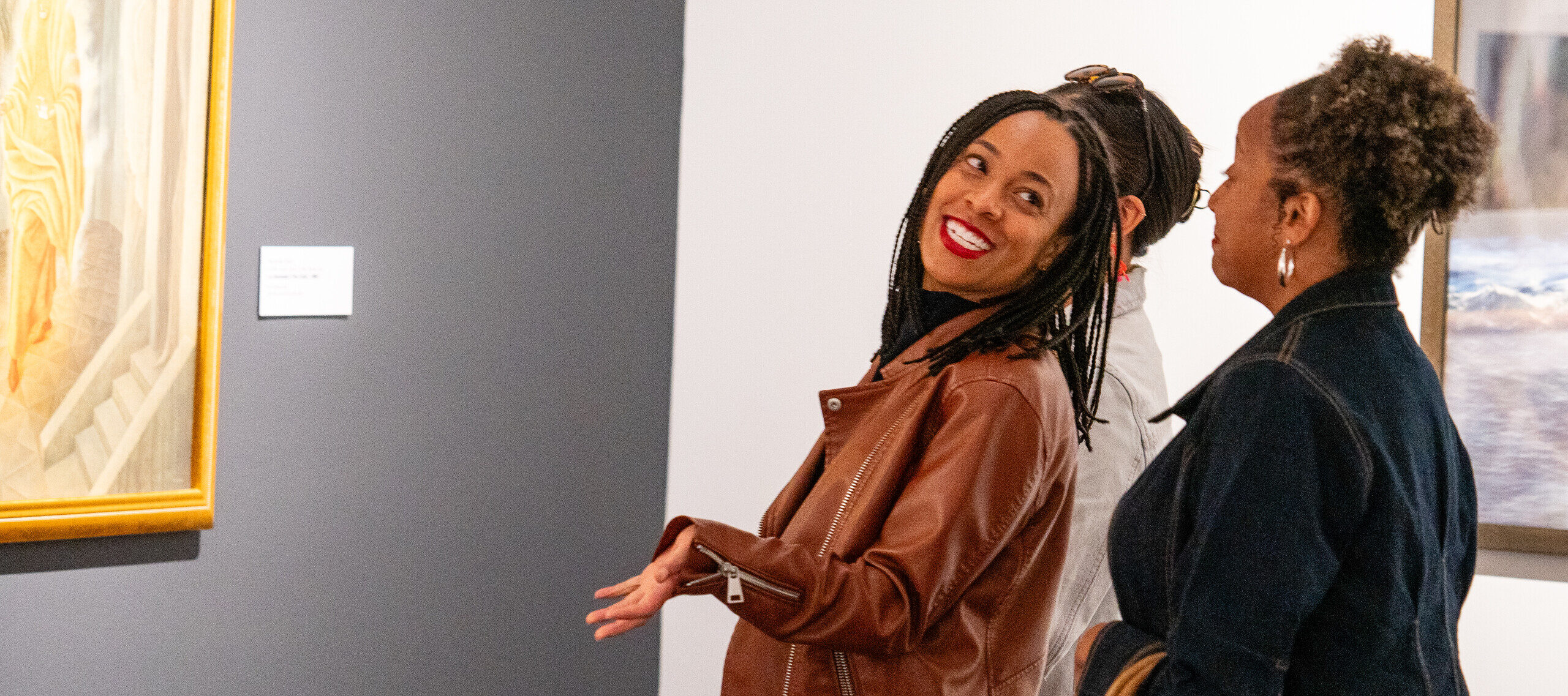Three people stand in an art gallery, engaging with a painting on the wall. One woman in a brown leather jacket gestures and smiles, while the others listen. They appear to be enjoying the exhibit.