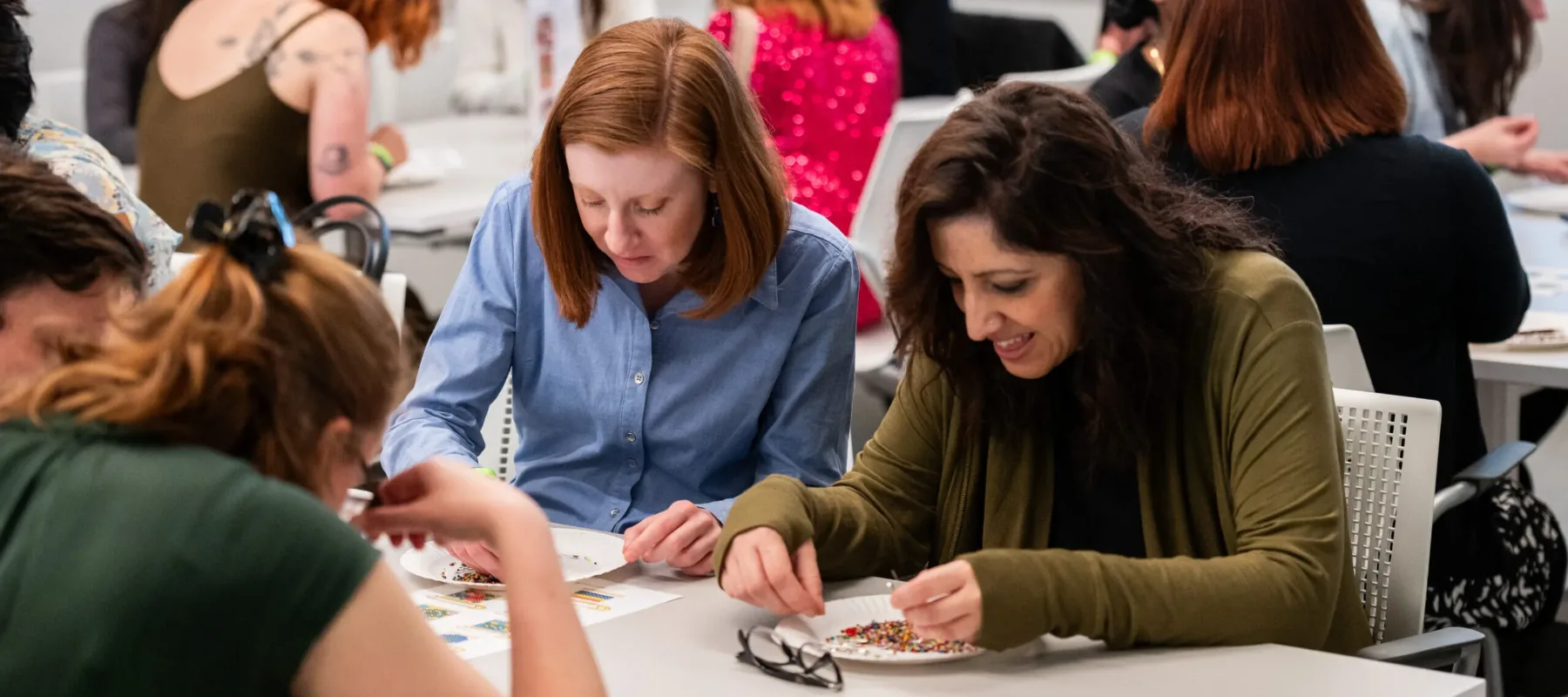Numerous people sit at white tables, sorting through white, paper plates with multicolored breads and safety pins.
