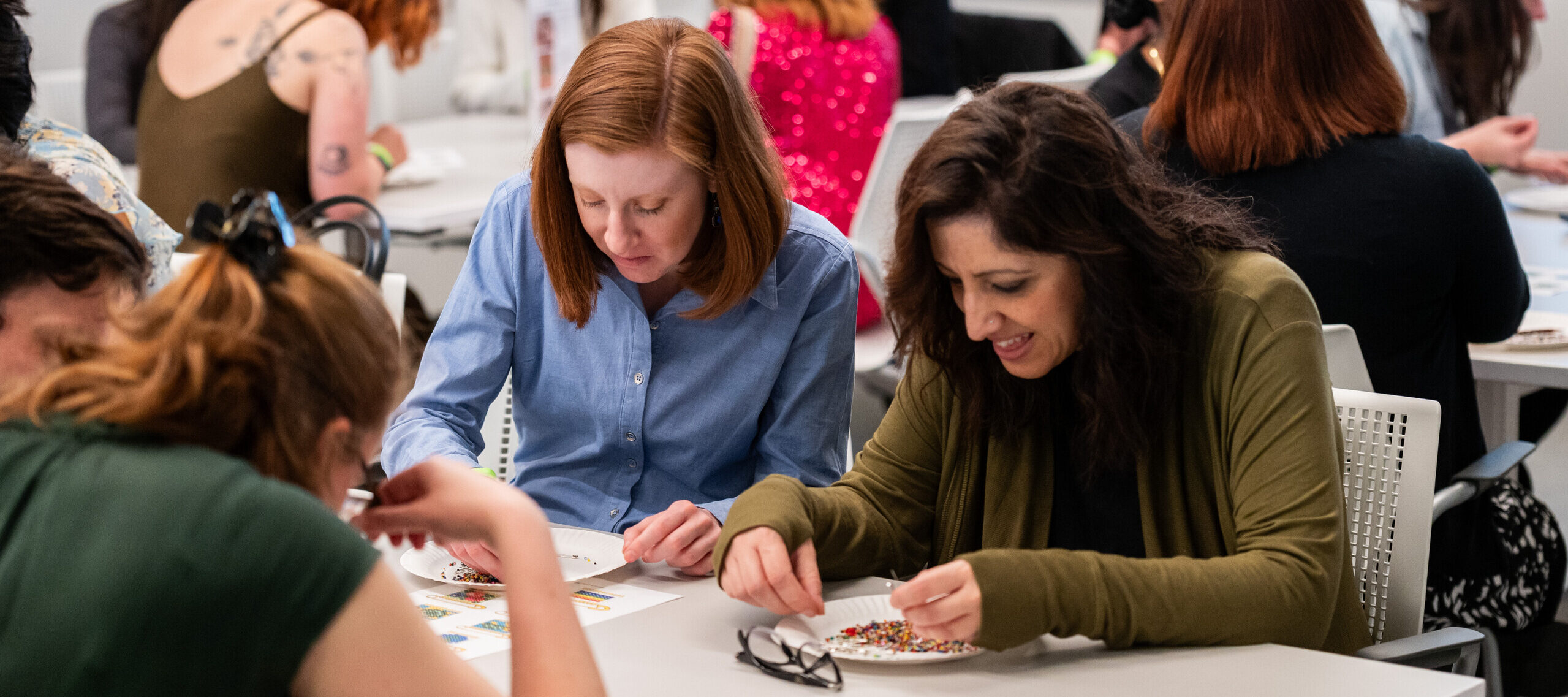 Numerous people sit at white tables, sorting through white, paper plates with multicolored breads and safety pins.