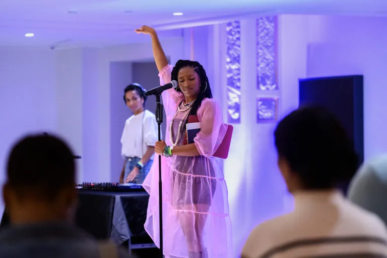 A woman in a pink sheer dress holds her hand up and performs into a microphone with a DJ behind her.