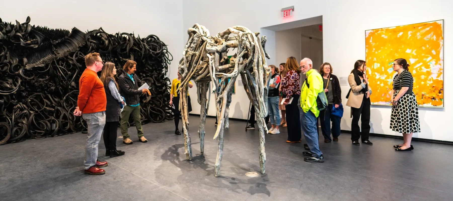 View of a gallery. Several people are standing in the space, viewing the artworks. A large, yellow painting hangs on the right wall, an even bigger sculpture made of rubber tires is on the left wall, and a sculpture made of branches stands in the middle of the room.