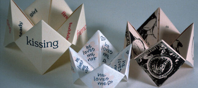 Three handmade paper fortune tellers, one with handwritten text, one with black ink illustrations, and one partially visible with printed text, are displayed on a pink background.