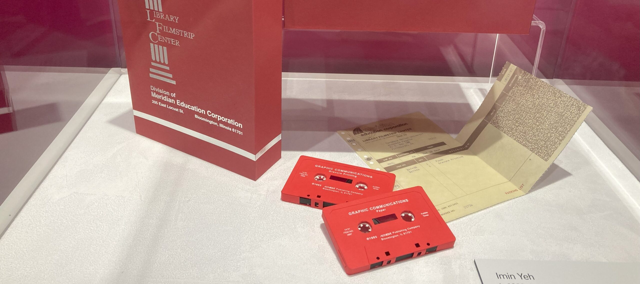 Two red cassette tapes and a printed receipt accompanied by two red film canisters and a gray book the size of a cassette tape with the title "Paper Paper Film" embossed on its cover in a red storage box with a lid that reads "Education Corporation."