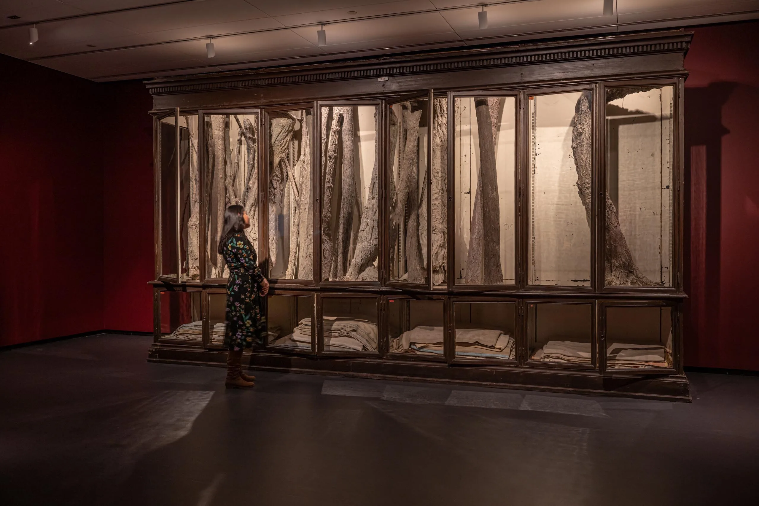 A woman in a black dress stands alone in a dimly lit art gallery space looking at a large art installation of a glass display case filled with trees.