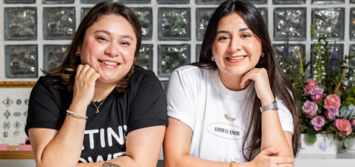 Two young women, both with straight dark brown hair, pose with their arms resting on a counter and one hand under their chin. They are under a fluorescent sign that says "JZD" in the design of a smiley face.