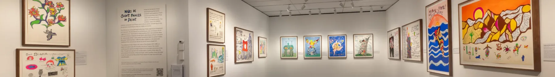 A modern art gallery features framed colorful drawings on white walls, track lighting overhead, and a black cushioned bench in the center of the dark floor. Informational panels are mounted between the artworks.