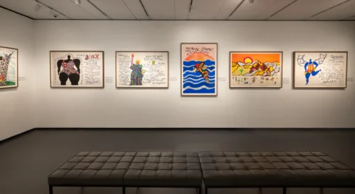 A contemporary art gallery with white walls displays colorful, framed artworks featuring handwritten text, patterns, and illustrations. A black cushioned bench sits in the room.