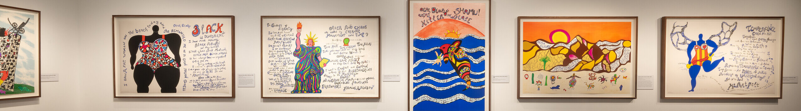 A contemporary art gallery with white walls displays colorful, framed artworks featuring handwritten text, patterns, and illustrations. A black cushioned bench sits in the room.