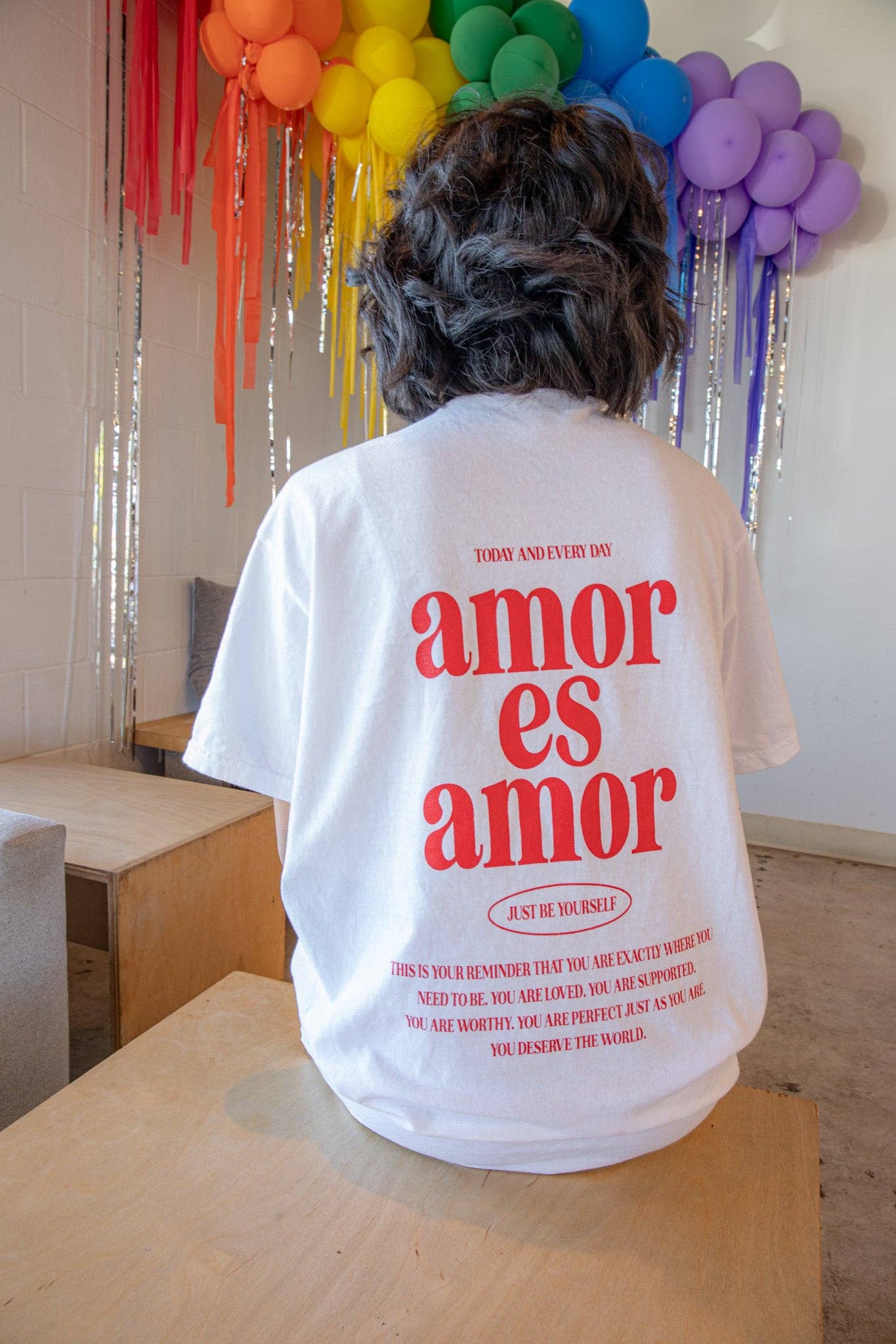 A person is photographed from the back, sitting on a table wearing a white T-shirt that reads "amos es amor" in large red font. More writing is visible in a smaller size below and above it. There is a display of rainbow balloons in front of the person.