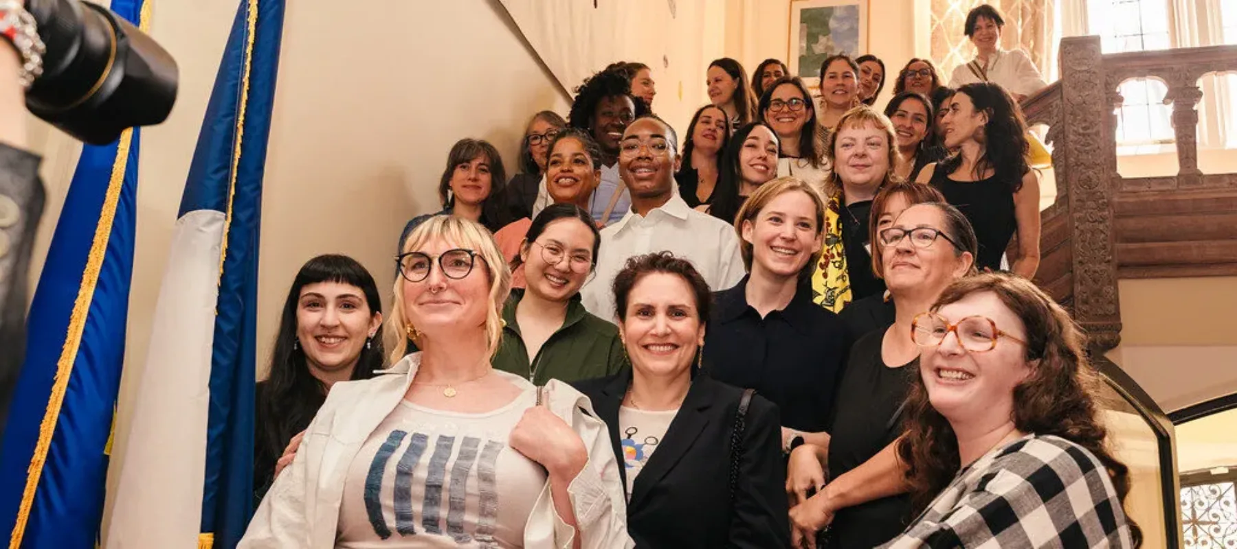A group of 28 women of varying ages and ethnicities stand on a marble staircase. There is a French flag on a pole to the left of the staircase, and the women are smiling down into the camera.