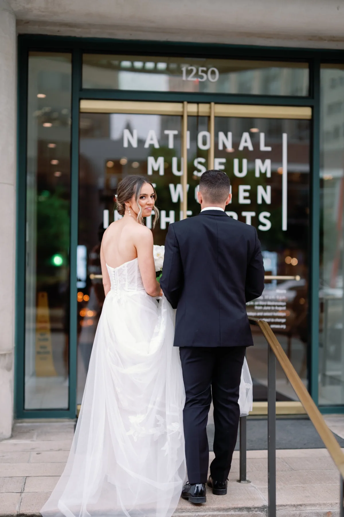 A bride and groom walk up the stairs towards the entrance to the National Museum of Women in the Arts. The bride looks over her shoulder at the photographer.