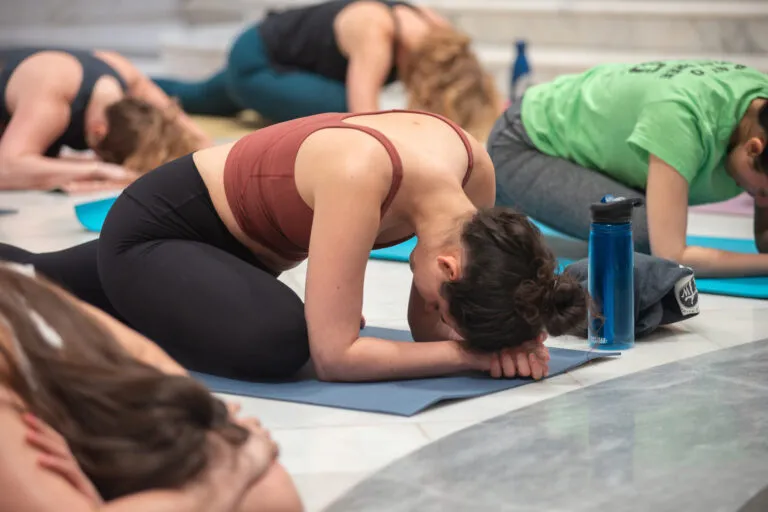A crowd participates in a yoga class in the Great Hall of the National Museum of Women in the Arts. They all wear athletic apparel and sit on their yoga mats in a "pigeon pose."