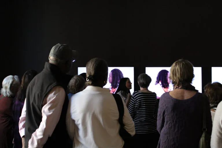 A woman is standing in front of a group next to a video screens. She is gesturing with her hands as she talks, engaging the group. She has medium-light skin tone, long hair, and is wearing a white shirt.