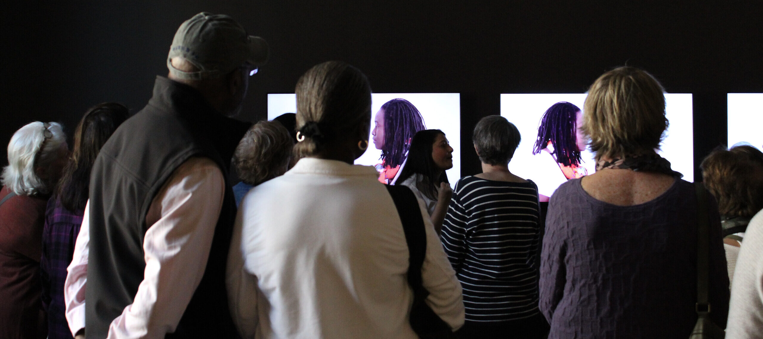 A woman is standing in front of a group next to a video screens. She is gesturing with her hands as she talks, engaging the group. She has medium-light skin tone, long hair, and is wearing a white shirt.