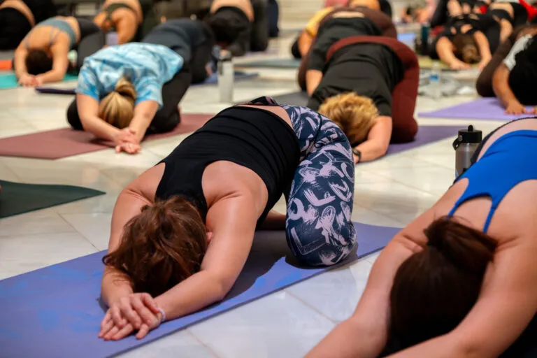 A crowd participates in a yoga class in the Great Hall of the National Museum of Women in the Arts. They all wear athletic apparel and lay down on their own yoga mats in a "child's pose" position.