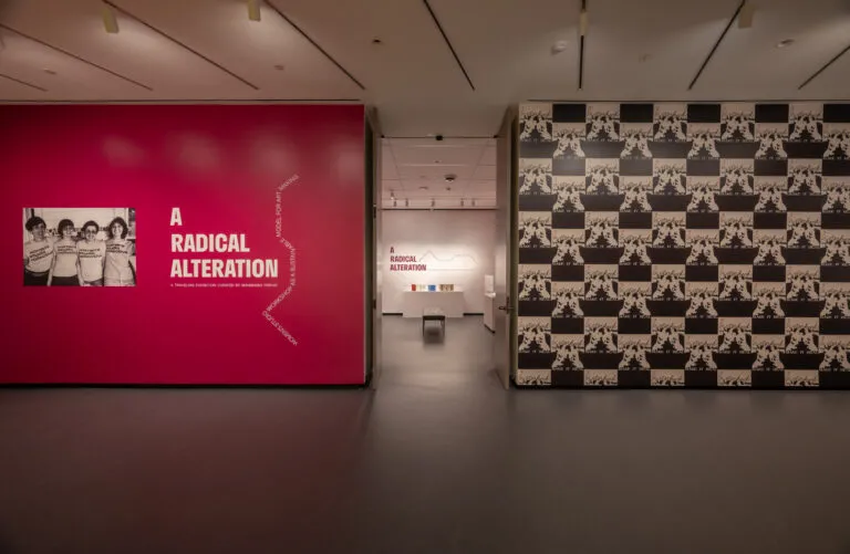 A gallery space with dark floors. On the left is a magenta wall that says "A Radical Alteration" with an enlarge black-and-white photo of four people posing shoulder-to-shoulder in t-shirts. On the right is a wall with a repeated black-and-white image in a large grid pattern.