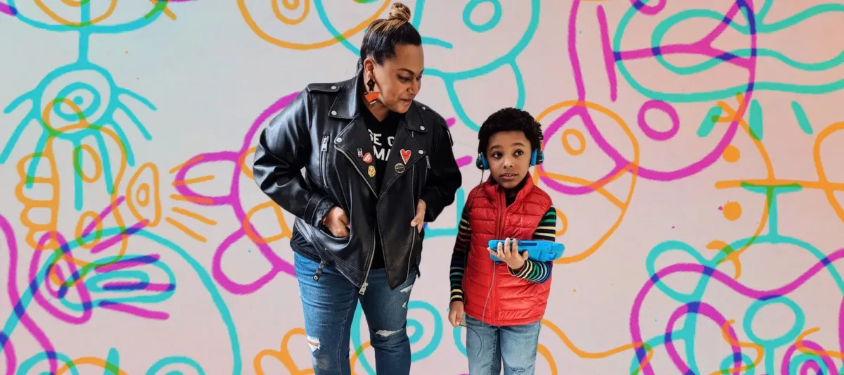 A woman with medium-dark skin tone stands next to a young boy with medium-dark skin tone. Their bodies have been edited atop a colorful, playful background of pinky, blue, and yellow shapes. The boy holds a digital bad and wears headphones.