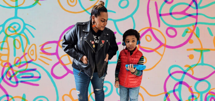 A woman with medium-dark skin tone stands next to a young boy with medium-dark skin tone. Their bodies have been edited atop a colorful, playful background of pinky, blue, and yellow shapes. The boy holds a digital bad and wears headphones.