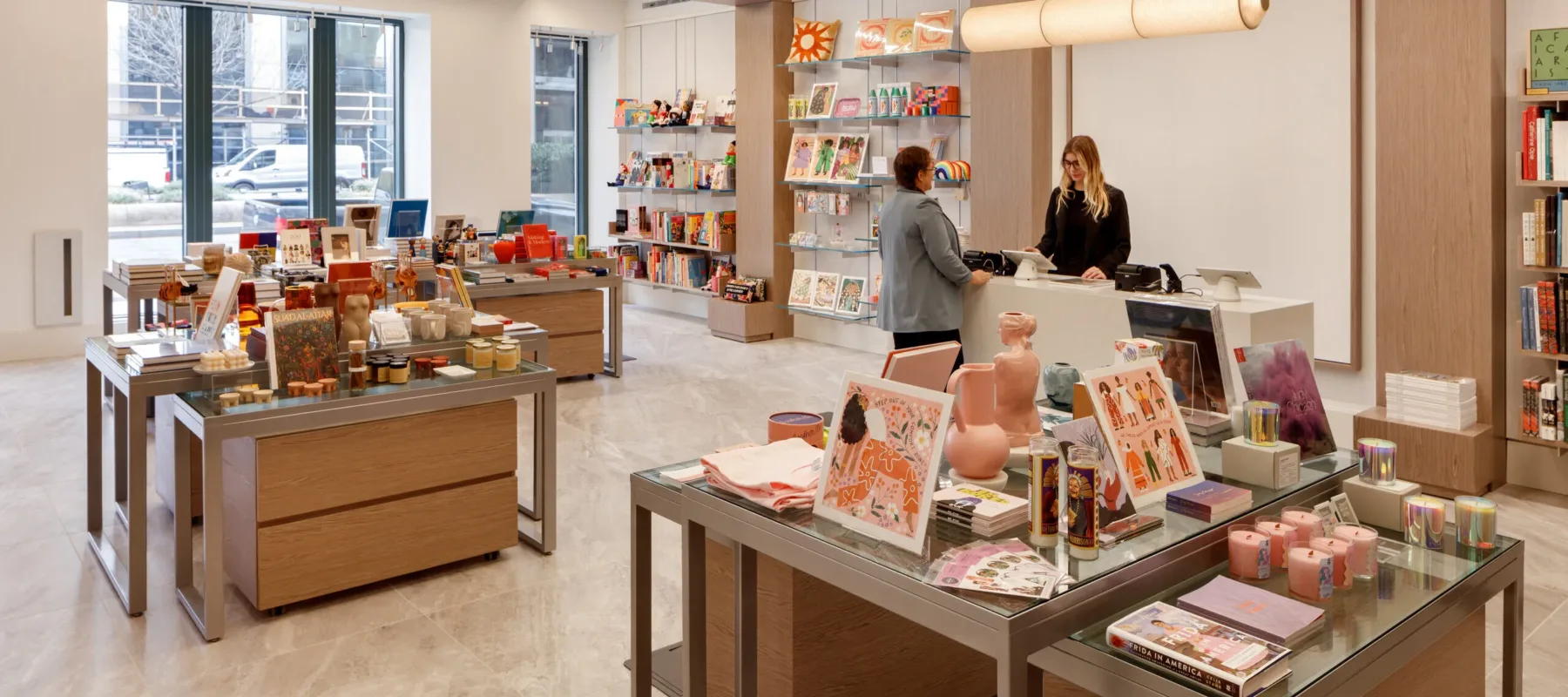 A museum gift shop is photographed at a wide angle. A colorful assortment of books, candles, textiles, and other small objects cover tables and shelves on the wall. A shopper is paying for items at a checkout table.